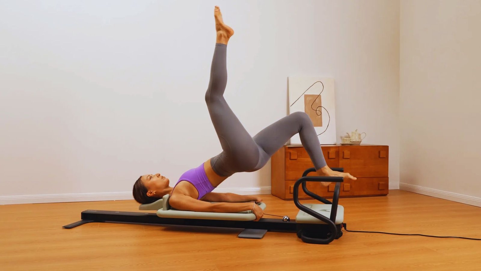 A woman doing a Pilates bridge on a reformer machine with one leg extended straight up.