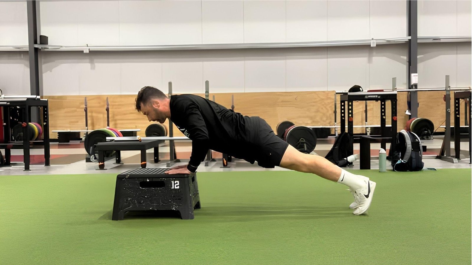 ** Man in black doing incline push-ups on plyo box in spacious gym with green turf floor.