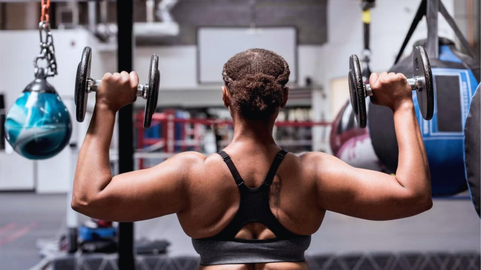 Muscular woman from behind does dumbbell shoulder press in boxing gym wearing black sports bra, punching bags hanging nearby.