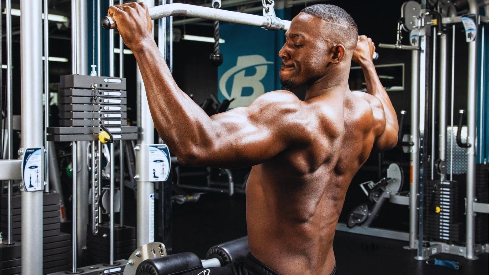 Shirtless muscular man performs lat pulldown on cable machine showing defined back muscles with gym equipment in background.