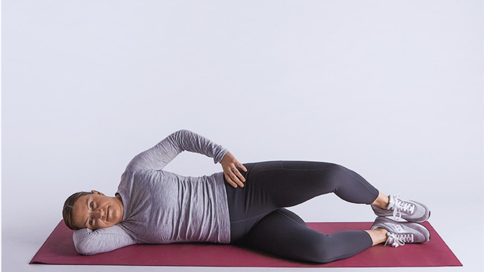 Woman in gray long-sleeve top and black leggings lies on side doing clamshell exercise on maroon mat, hand on hip.