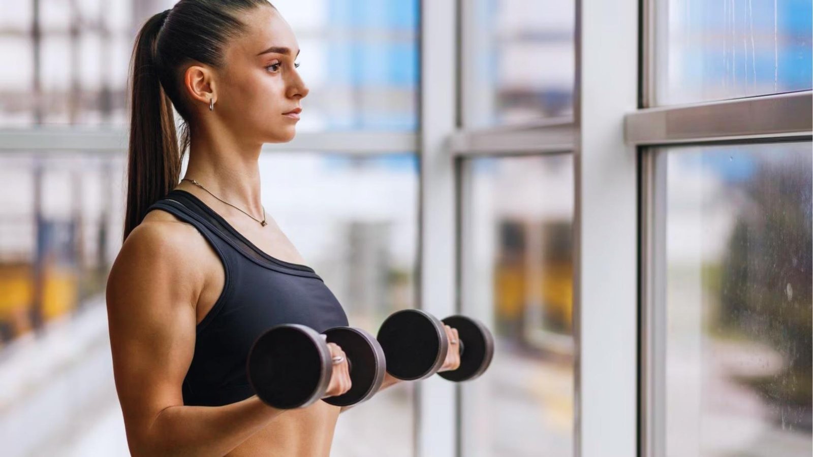 Focused woman with high ponytail performs dumbbell bicep curls in black sports bra near large gym windows with city view.