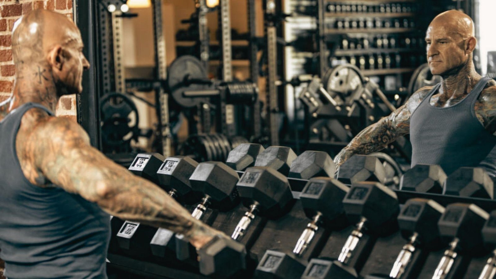 A man with visible tattoos stands before a mirror, reflecting on his image with a contemplative expression.