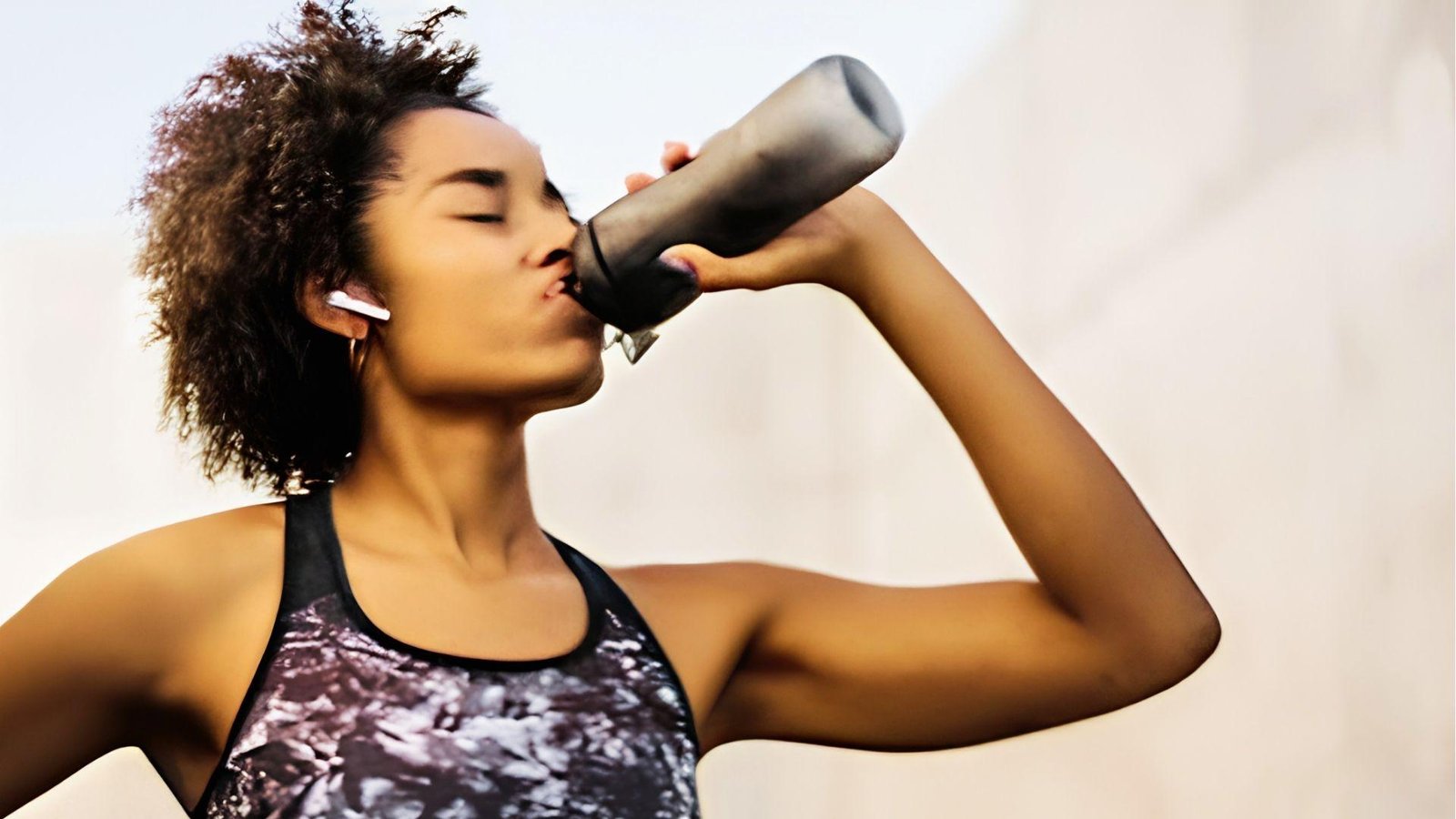 Woman with curly hair drinking pre workout from black shaker bottle outdoors