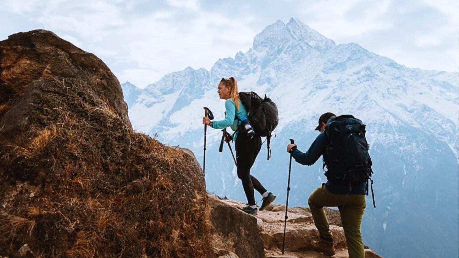 Two hikers with trekking poles and backpacks climb steep rocky mountain trail with snowcapped peaks and bright blue sky behind.