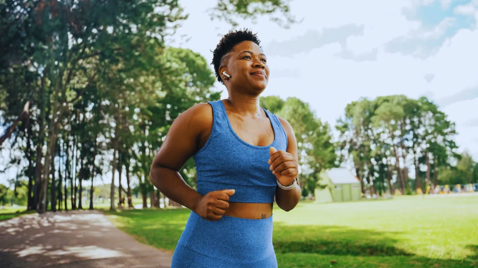 An athletic woman with a wireless earbud smiles while jogging outdoors on a sunny day.