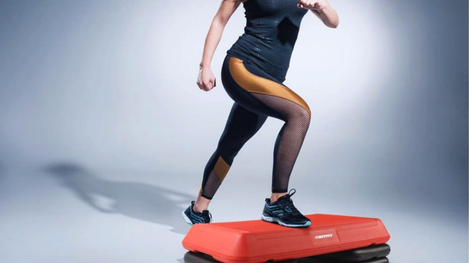 Athletic woman in black tank top and gold-mesh leggings steps onto red aerobic platform in studio, mid-motion, wearing black athletic shoes.