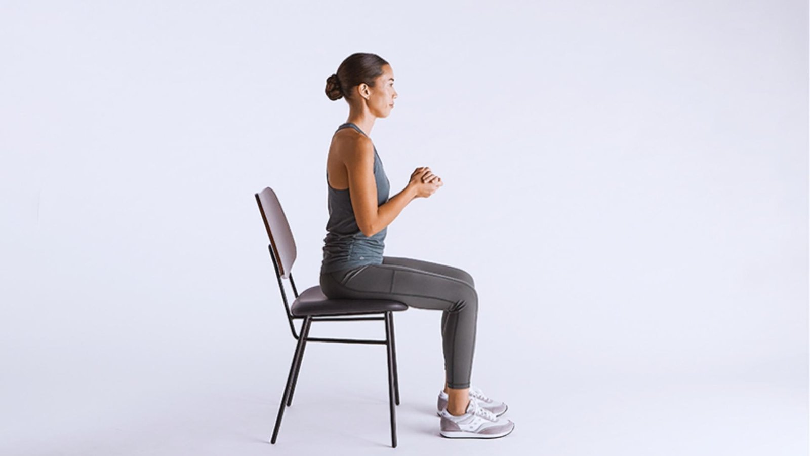 Woman with bun in gray tank top and leggings sits upright on black chair, hands clasped, feet flat on white studio floor.
