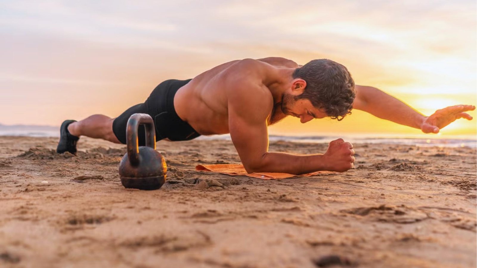 Shirtless muscular man performs plank with arm extended on sandy beach at sunset, kettlebell beside orange towel.