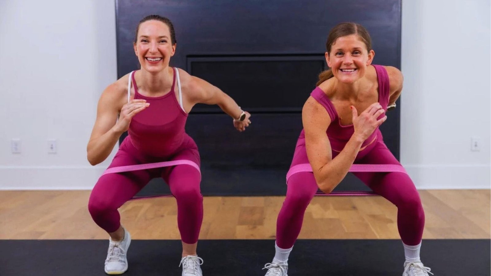 Two smiling women in matching magenta workout sets do banded squats on black mat, pink resistance bands above knees.