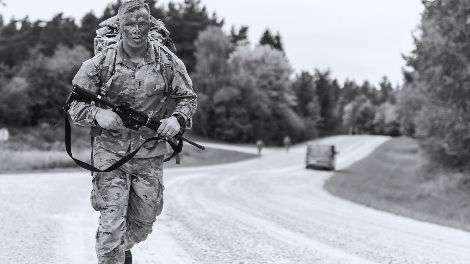 Black and white photo of U.S. soldier in camouflage uniform with face paint and rucksack, running on gravel road while carrying rifle.