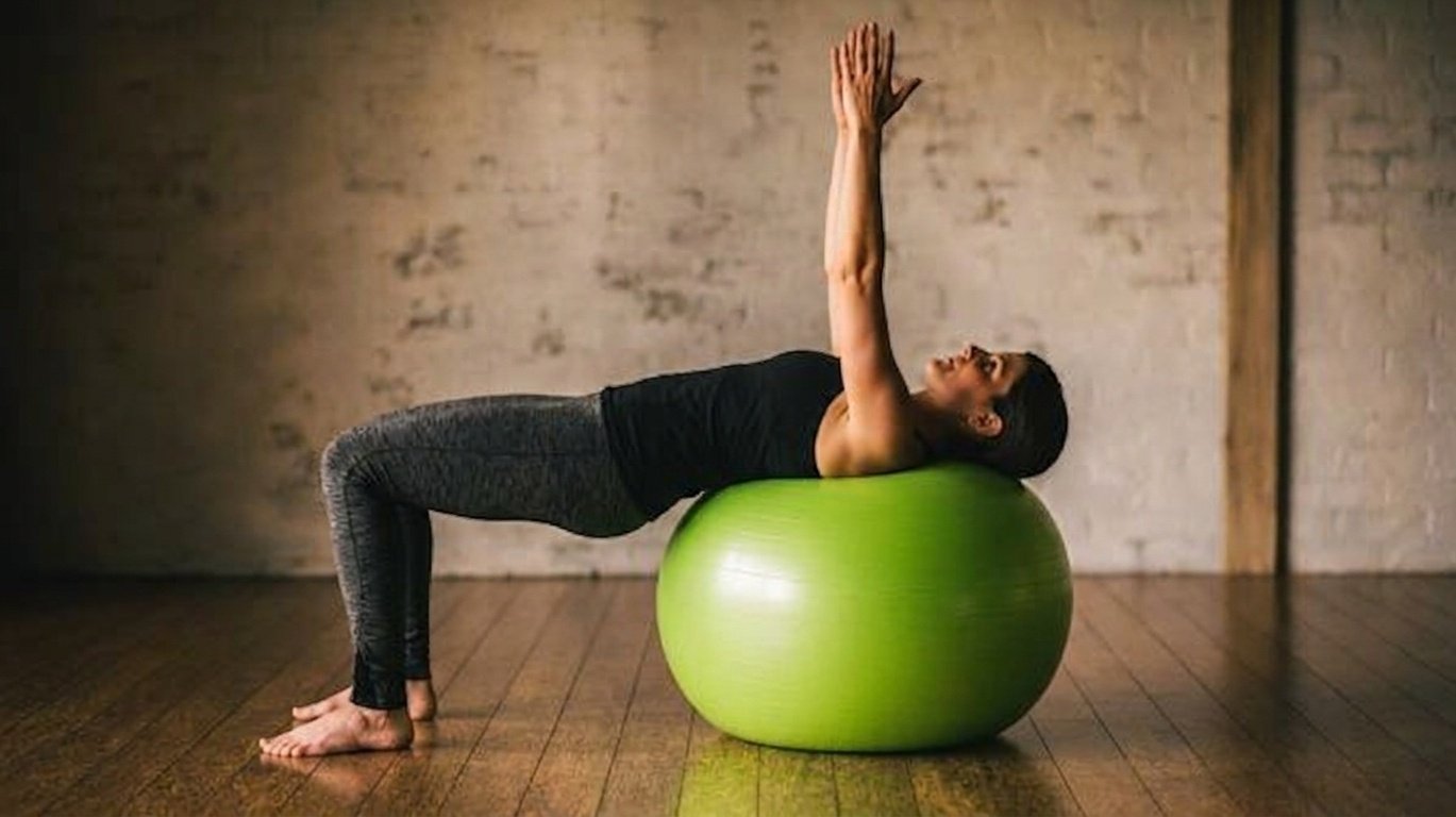 Woman doing the ball roller press while lying face up on the stability wall and her arms kept straight.