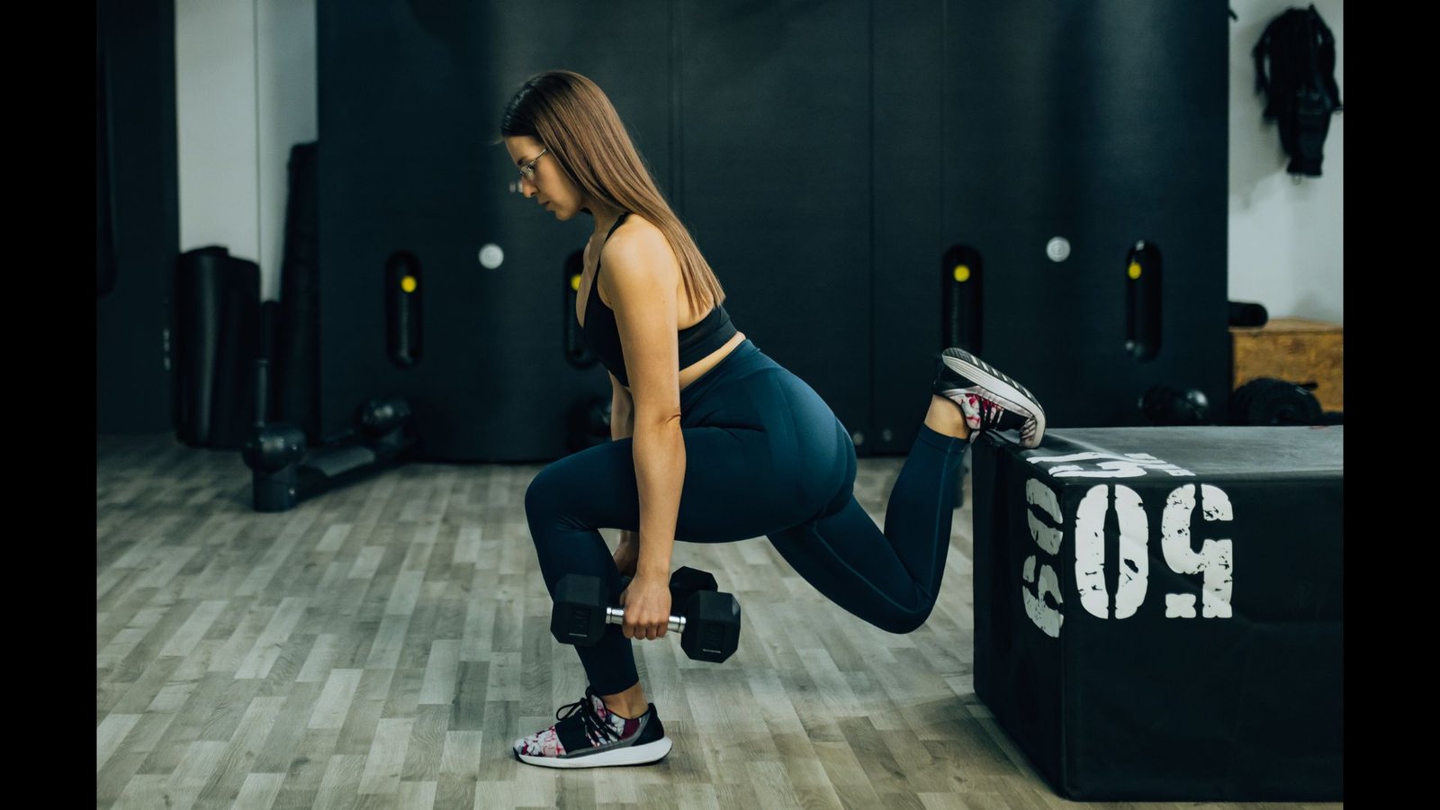 A woman in athletic wear holds a deep Bulgarian split squat with a dumbbell, her back foot resting on a gym box.