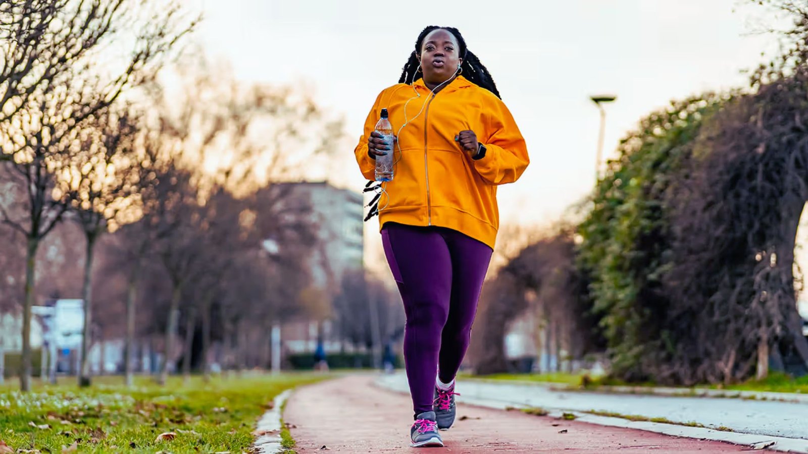 An athletic woman in an orange jacket and purple leggings walks briskly along an outdoor path.