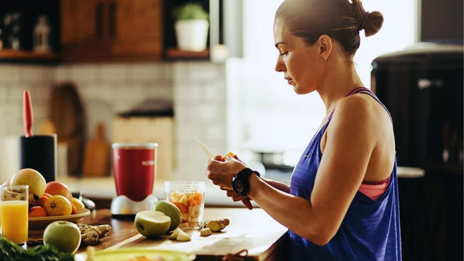 Woman in blue tank top slices orange fruit in sunny kitchen with blender, apples, ginger, and juice on countertop.