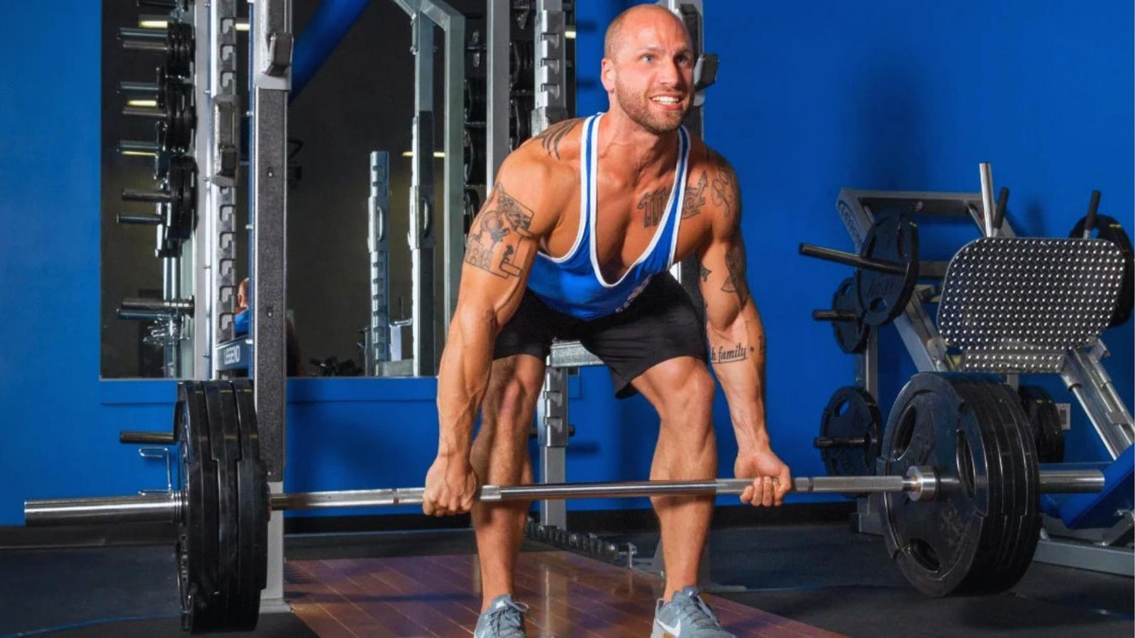 Tattooed man in blue tank top and black shorts does barbell deadlift with heavy plates in bright blue gym.