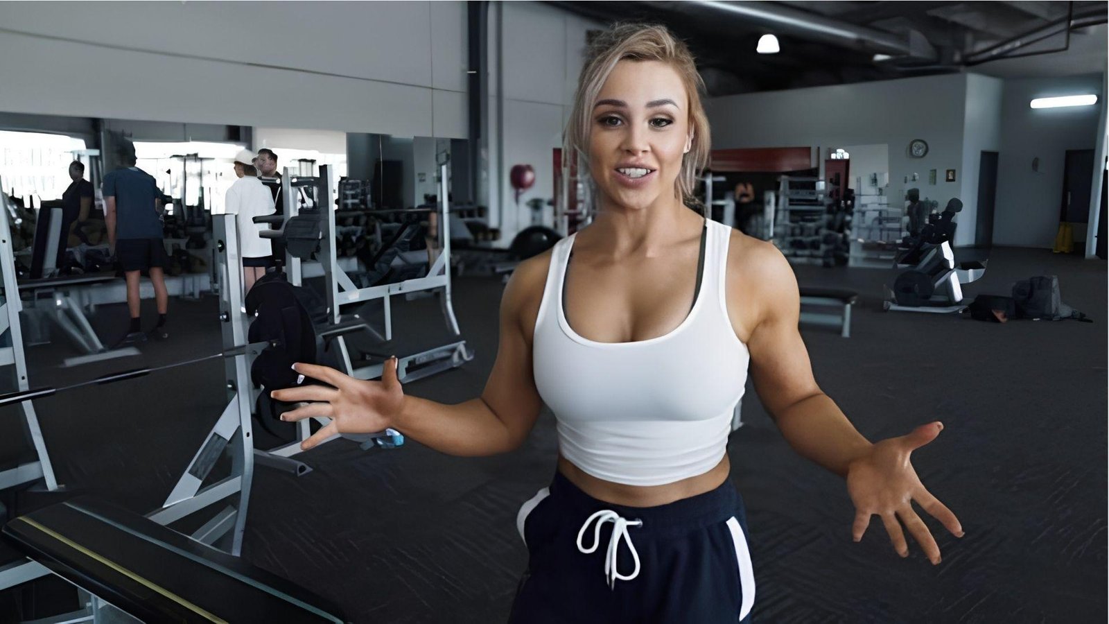 Fit blonde woman in white crop top explaining chest workout in modern gym with equipment behind.