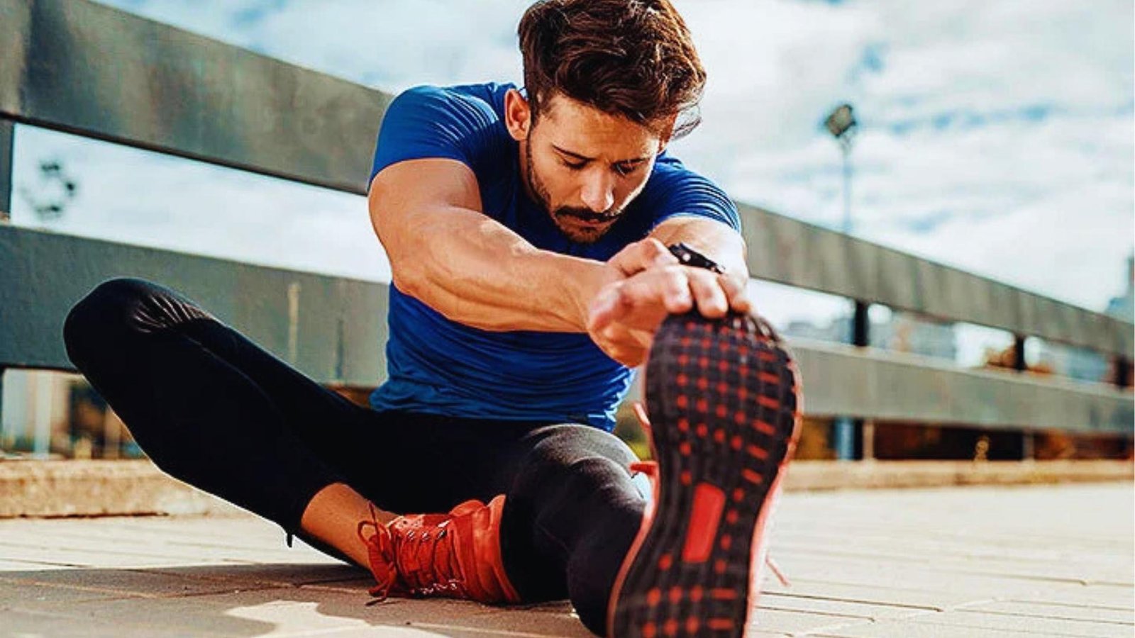 Man in blue shirt and black leggings does seated hamstring stretch on outdoor pavement, reaching for red running shoe sole.