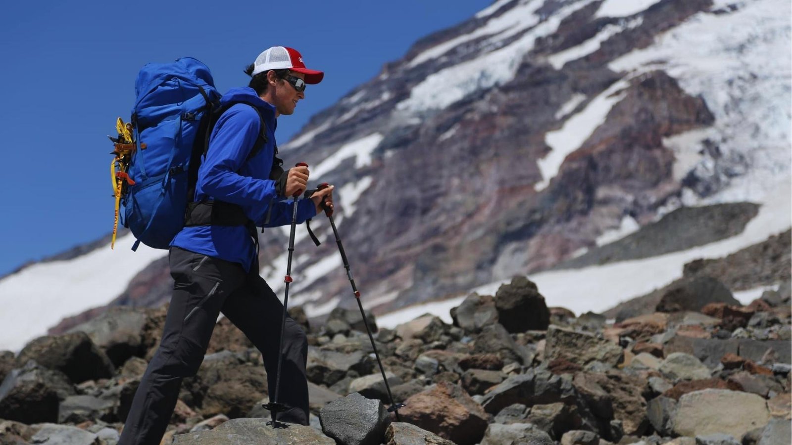 Hiker in blue jacket and red cap with trekking poles and blue backpack climbs rocky mountain slope with snowcapped peaks behind.