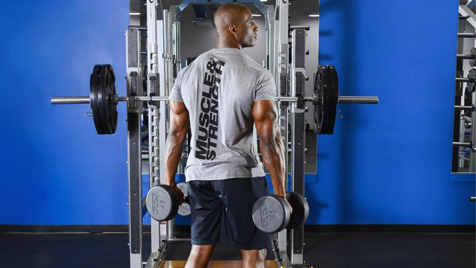 Muscular man in gray "Muscle & Strength" shirt holds heavy dumbbells in blue gym with loaded barbell rack behind.