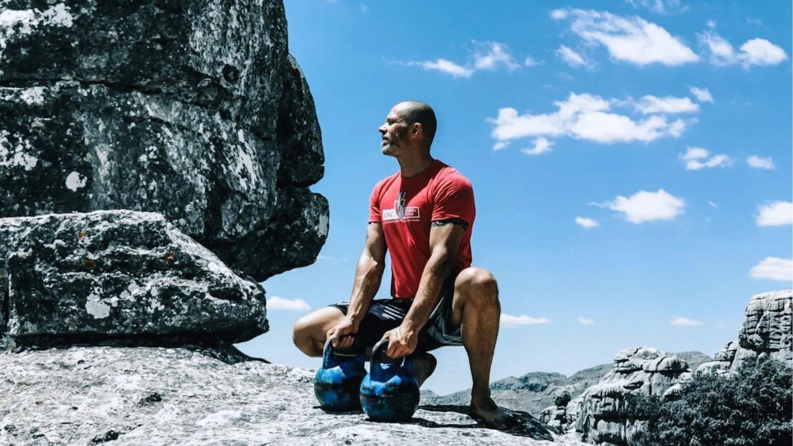 Bald man in red shirt squats holding two blue kettlebells on rocky mountain summit under bright blue sky with clouds.