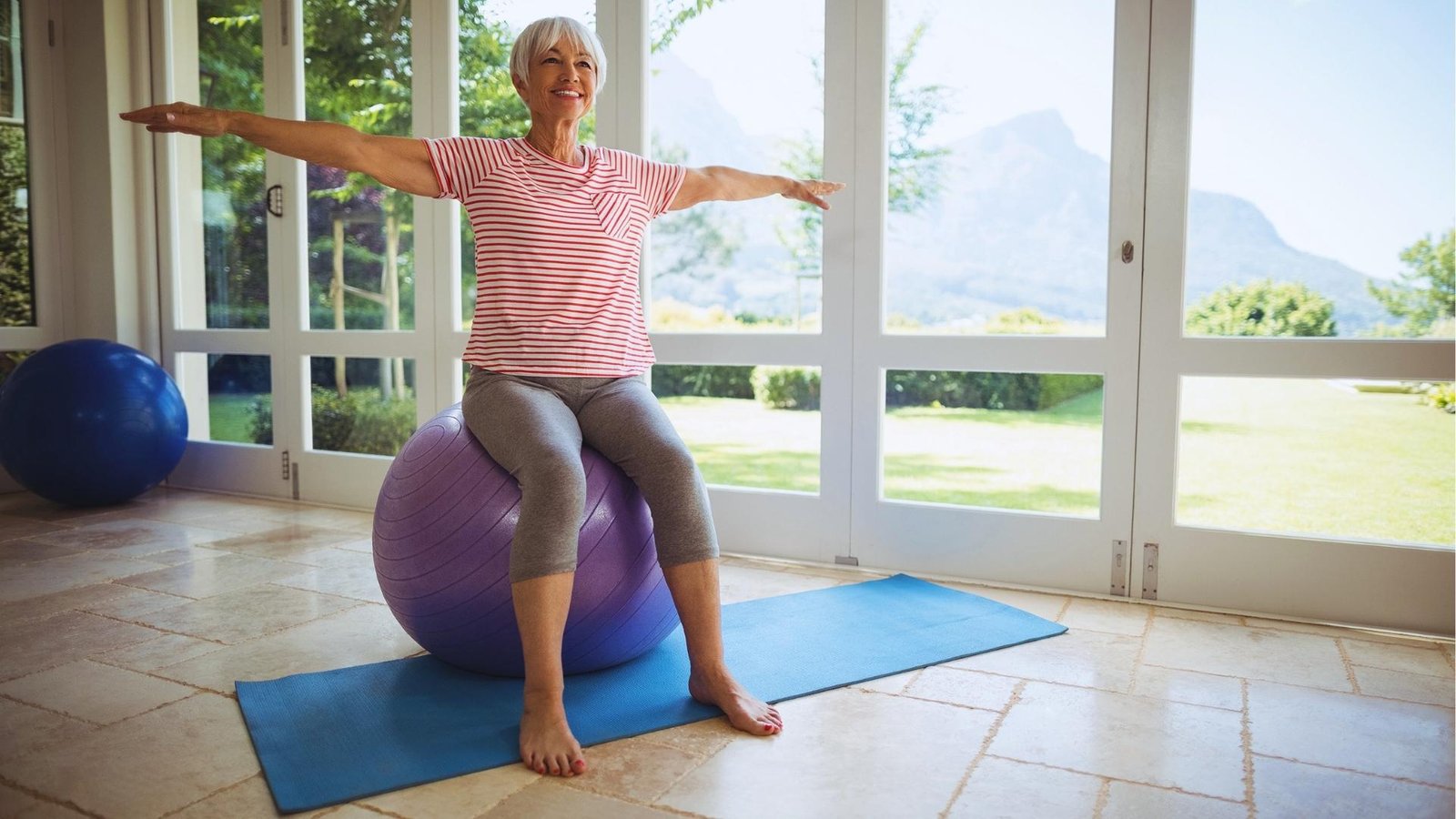 Older woman with short white hair sits smiling on a purple exercise ball, arms extended, balancing indoors with a mountain view behind.
