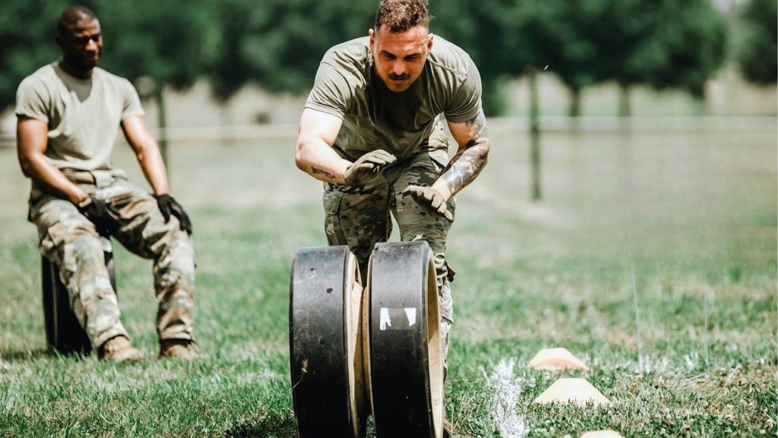 Soldier in camo pants and gloves pushes heavy weighted wheels across a grass field while another soldier watches from the background.