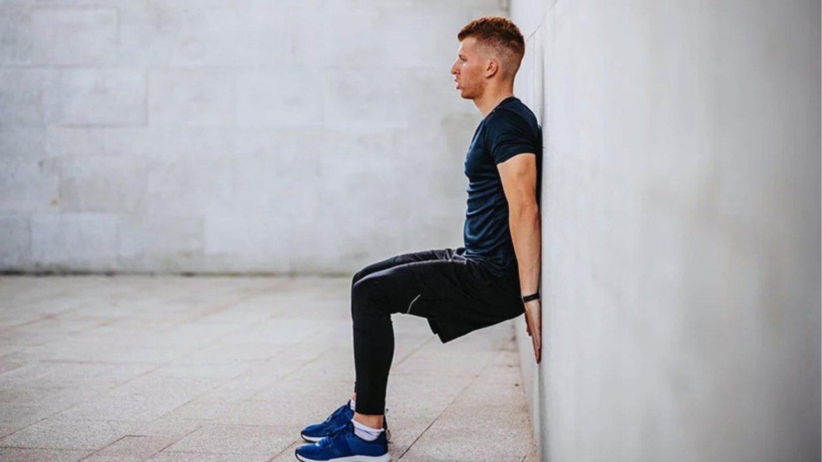 Man in black shirt and tights sits against concrete wall outdoors, knees bent 90 degrees, wearing blue shoes.