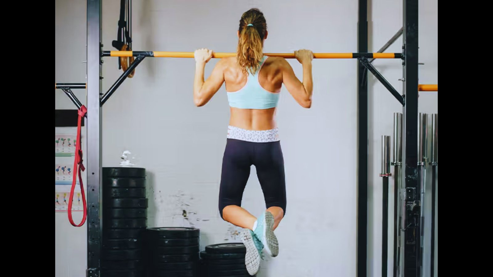 An athletic woman performs a pull-up, viewed from behind, gripping a yellow bar in a gym.