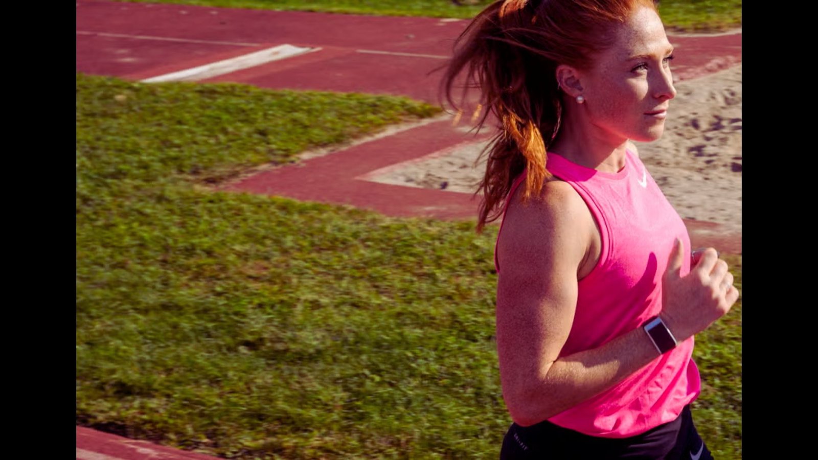 A woman with reddish hair in a pink tank top running outdoors on a sunny day.