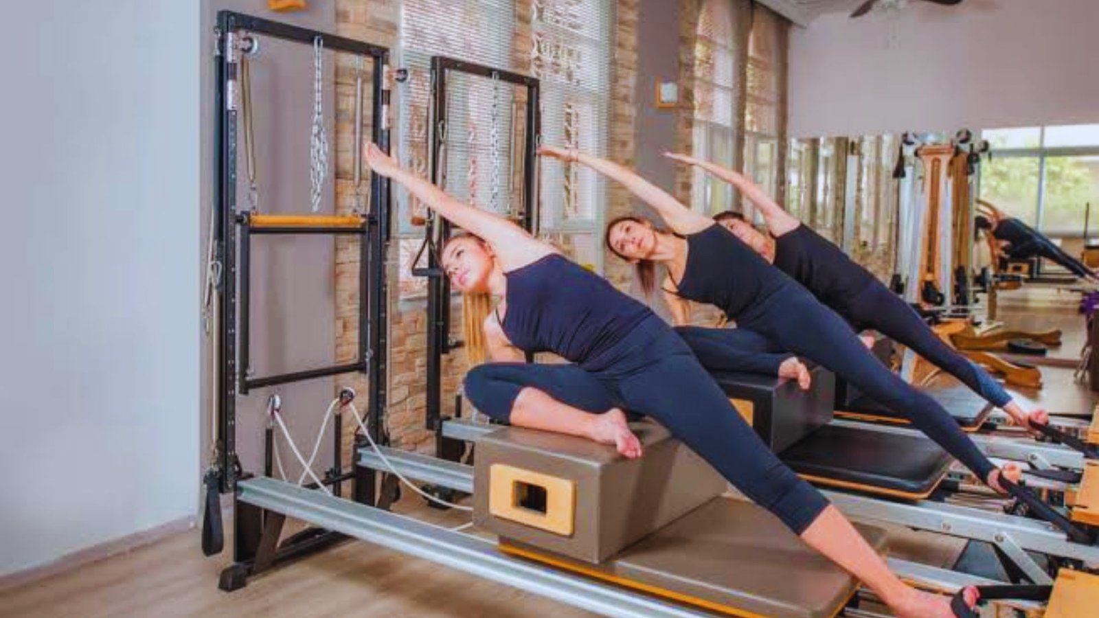 Three women doing a side plank exercise on Pilates reformer machines in a studio.