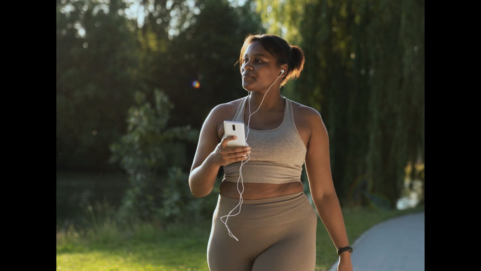 An athletic woman with earphones listens to her phone while walking outside during a sunny day.