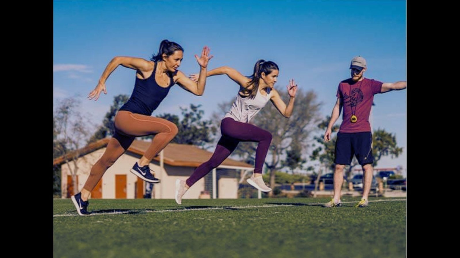 Two women perform jump squats outdoors on a sunny grass field while a trainer watches.