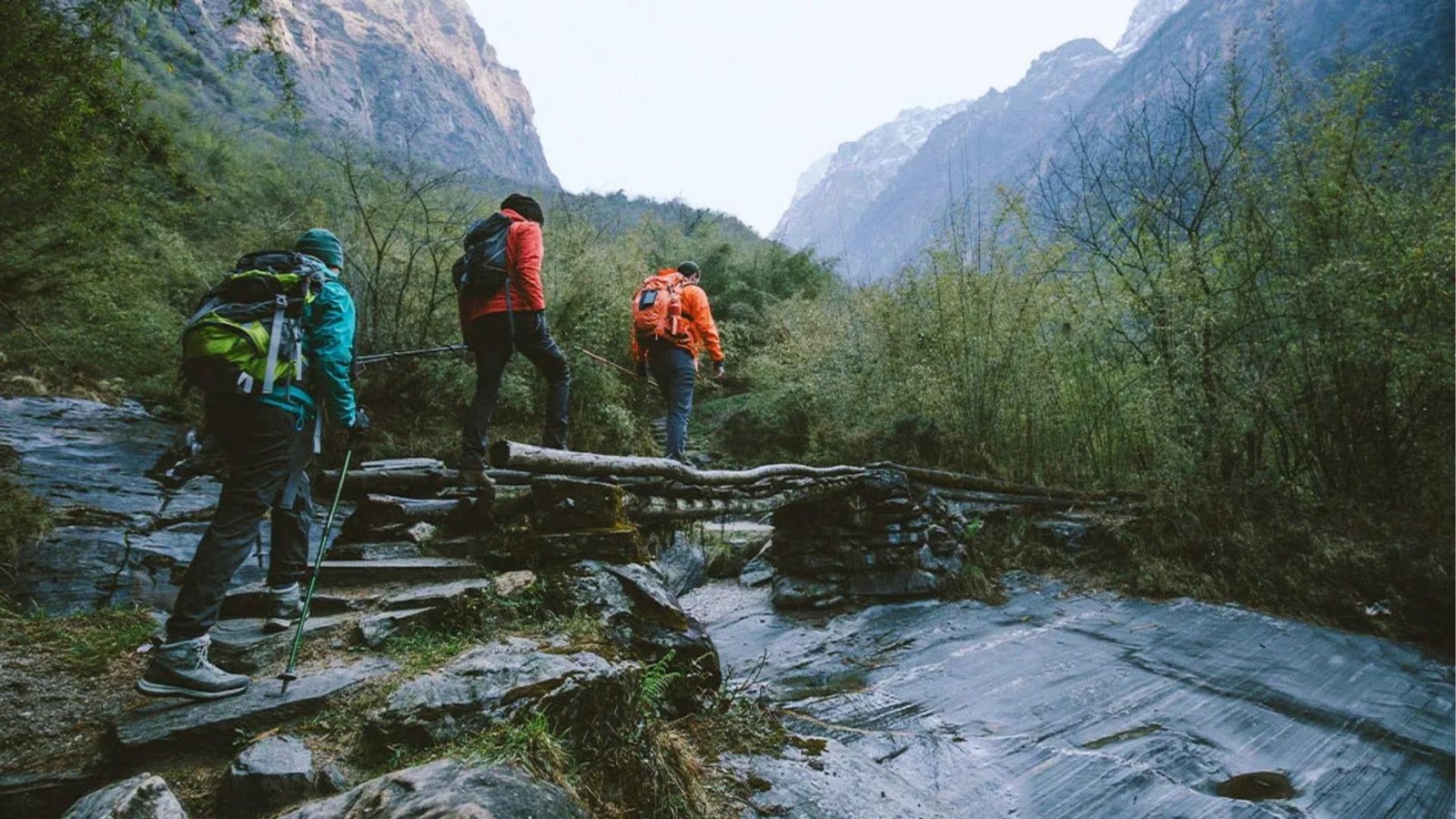 Three hikers with colorful backpacks cross a rustic log bridge over a stream in a lush mountain valley with steep cliffs surrounding.