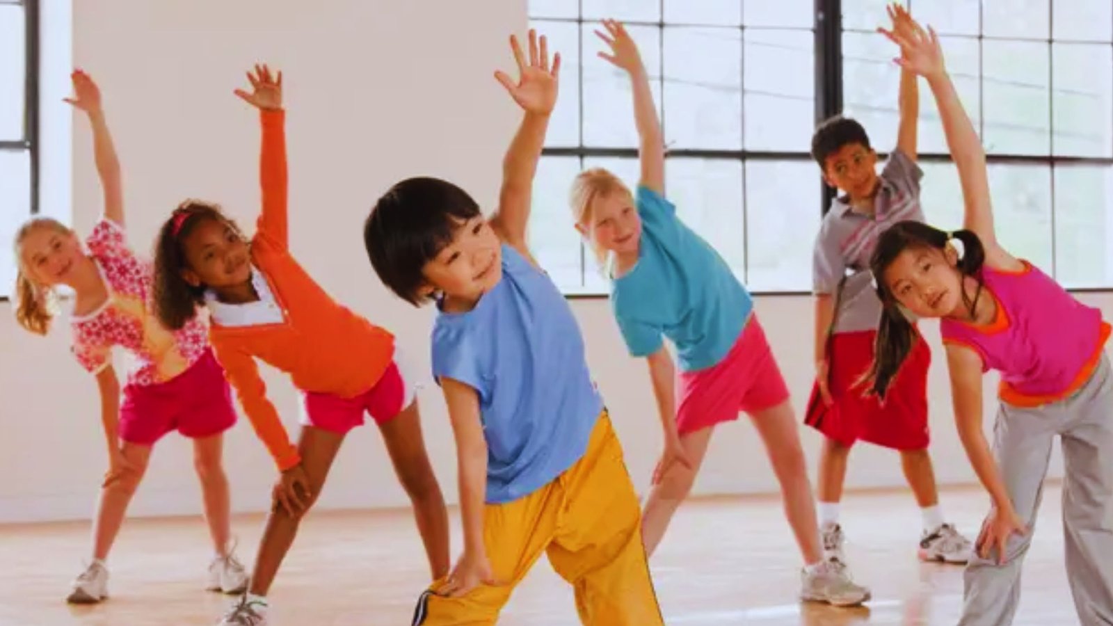 A cheerful group of children in sportswear smiling and exercising together in a bright, indoor room.