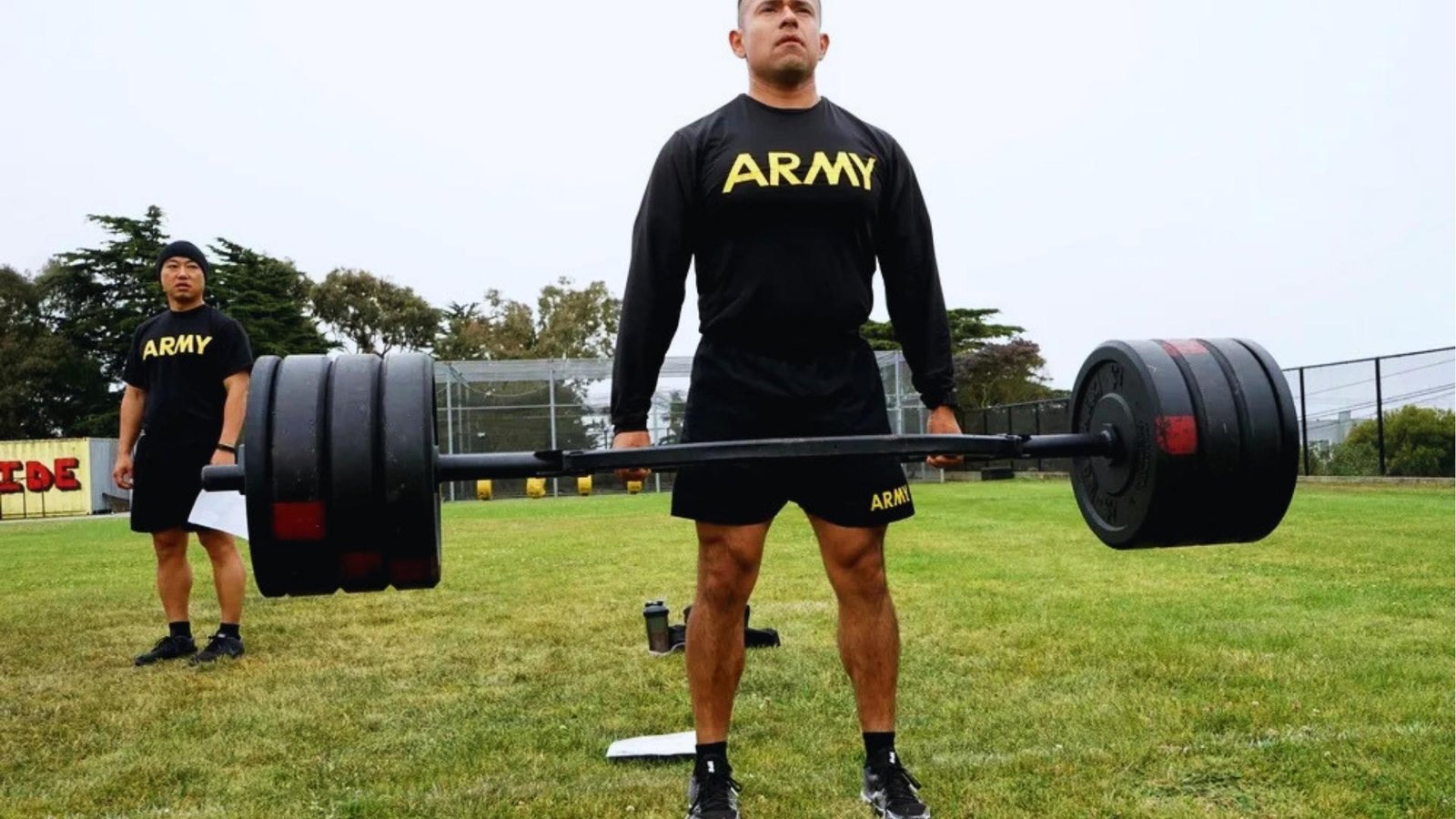 Soldier in "ARMY" shirt performs hex bar deadlift on grass field while another soldier watches with clipboard during assessment.