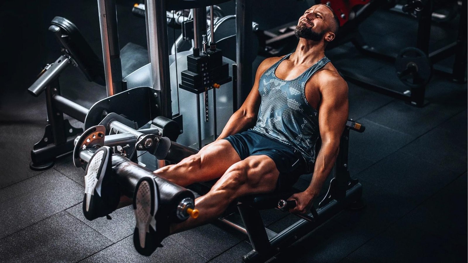 Muscular bearded man in patterned tank top strains while extending legs on leg extension machine in dark gym.